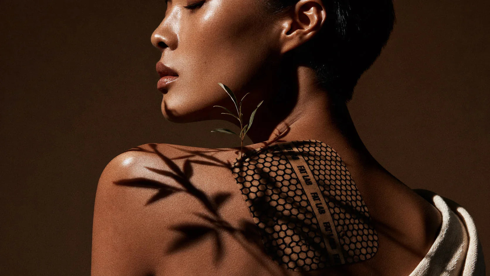 FATLAB skin tightening and toning body patch worn on a model’s shoulder with leaf shadows on a warm brown studio background